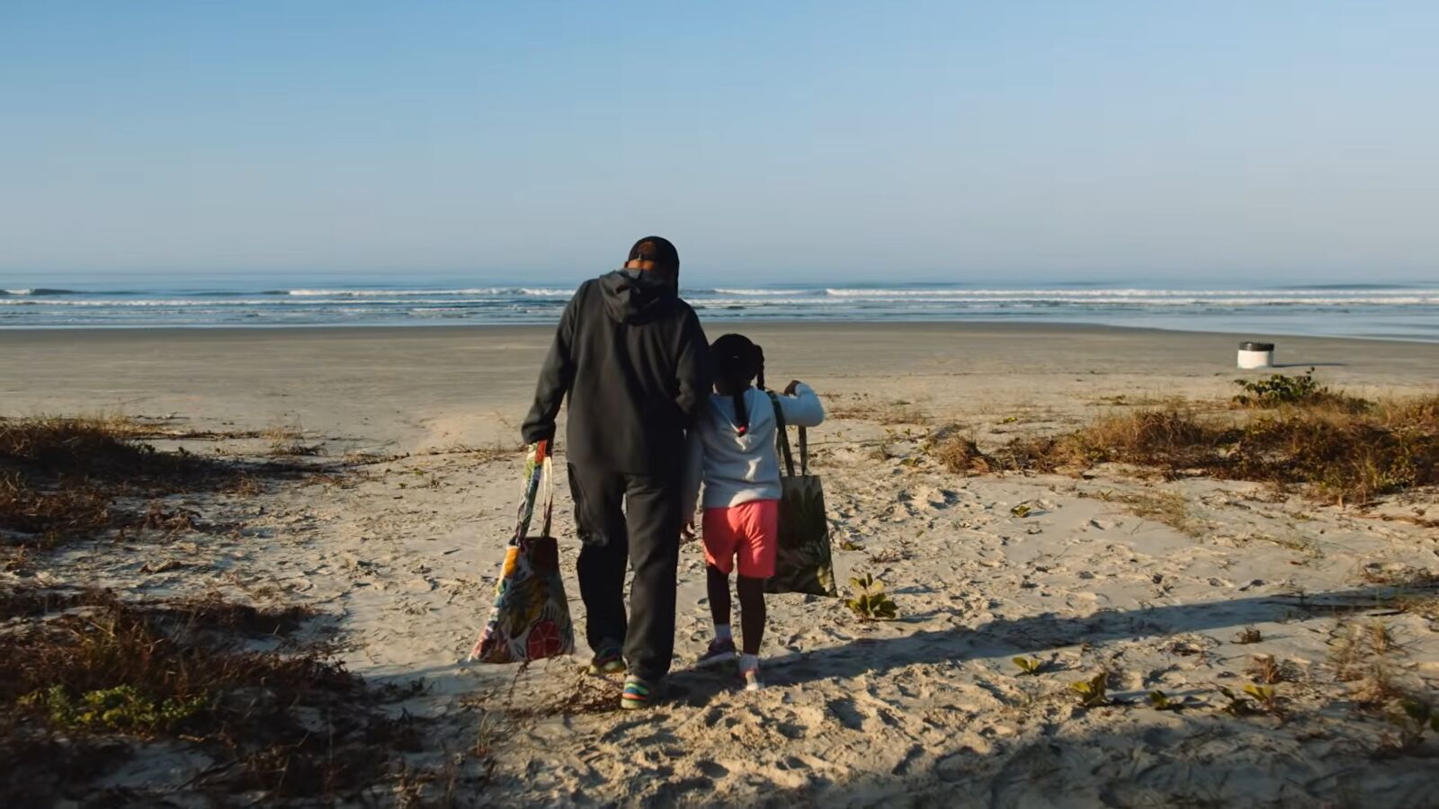 Adulto e criança caminhando juntos para o mar, numa praia vazia de manhã